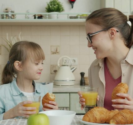 Young woman and adorable little girl are eating croissants and drinking juice talking at breakfast at home, modern interior is visible in background.
