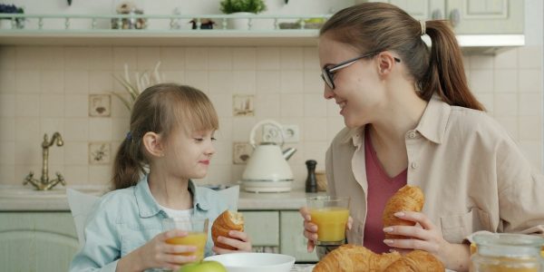 Young woman and adorable little girl are eating croissants and drinking juice talking at breakfast at home, modern interior is visible in background.