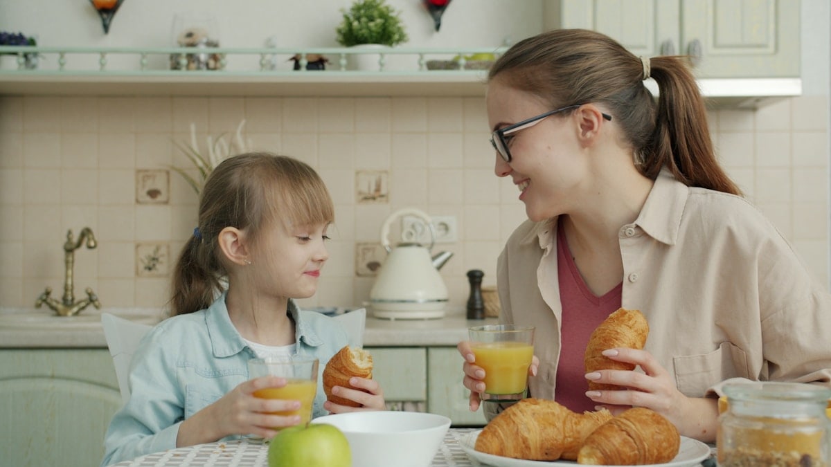 Young woman and adorable little girl are eating croissants and drinking juice talking at breakfast at home, modern interior is visible in background.
